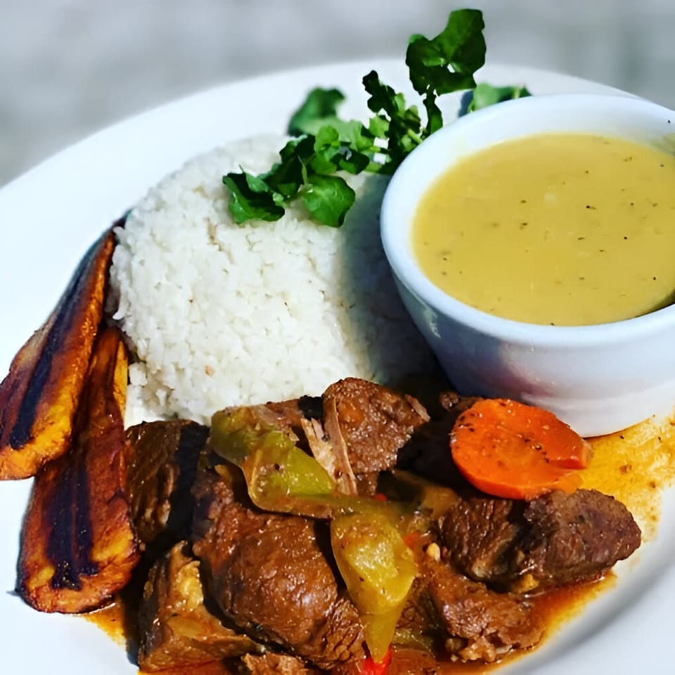 A Belizean Caribbean dish with stewed meat, rice, fried plantains, and a side of soup.